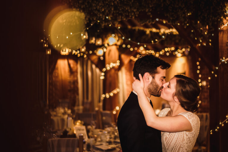 A wedding couple kissing to the backdrop of golden fairy lights in The Ferry House wedding barn at twilight