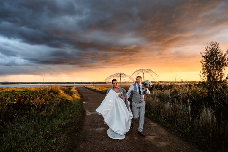 A wedding couples walking away from the Swale estuary at sunset with umbrellas in hand.