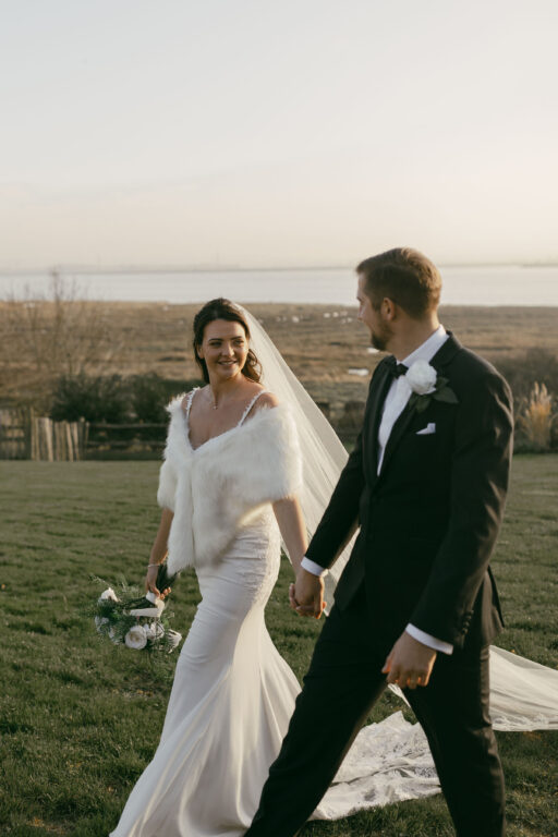 A couple walking through the frosty gardens of winter wedding venue The Ferry House in Kent with the Swale estuary in the background.