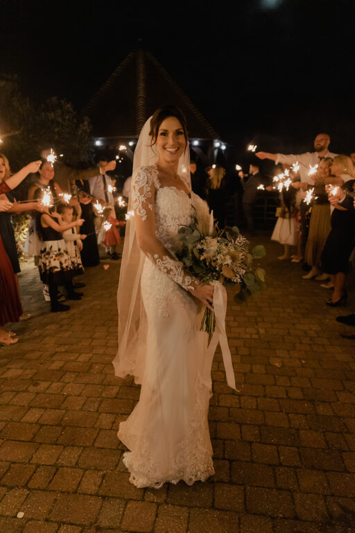 A bride posing with guests holding sparklers in the background on her wedding day at the winter wedding venue, The Ferry House in Kent.