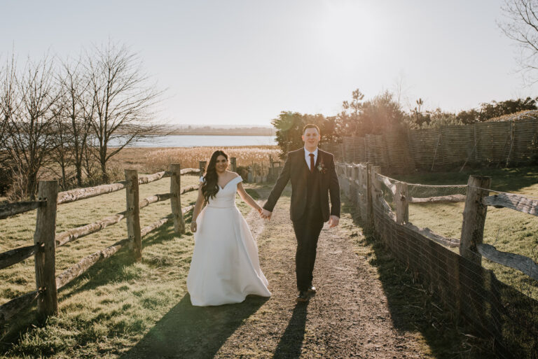 A bride and groom walking up from the Swale estuary at Kent barn wedding venue, The Ferry House, with the sun shining behind them.