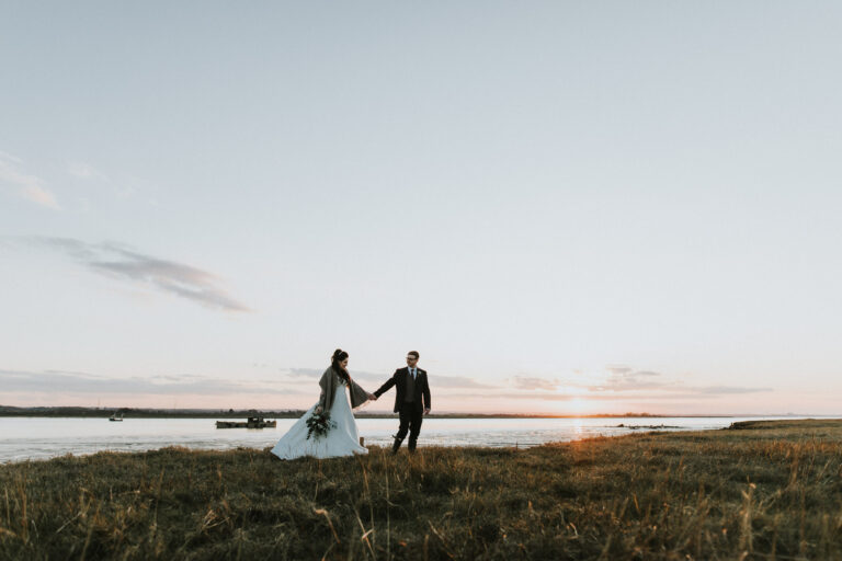A couple walking hand in hand along the Swale estuary at sunset on their wedding day at The Ferry House waterside wedding venue in Kent.